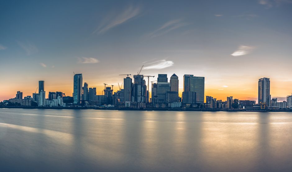 A wide panoramic view of the Canary Wharf skyline at sunset, featuring modern skyscrapers and office buildings constructed from glass and steel, with some under construction using cranes visible. The