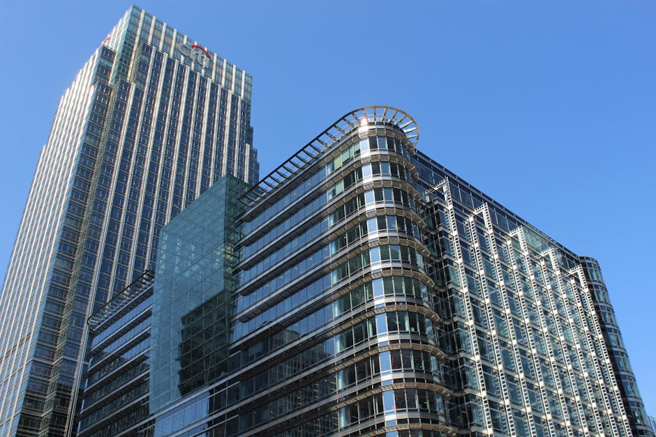 A wide panoramic view of the Canary Wharf skyline at sunset, featuring modern skyscrapers and office buildings constructed from glass and steel, with some under construction using cranes visible. The