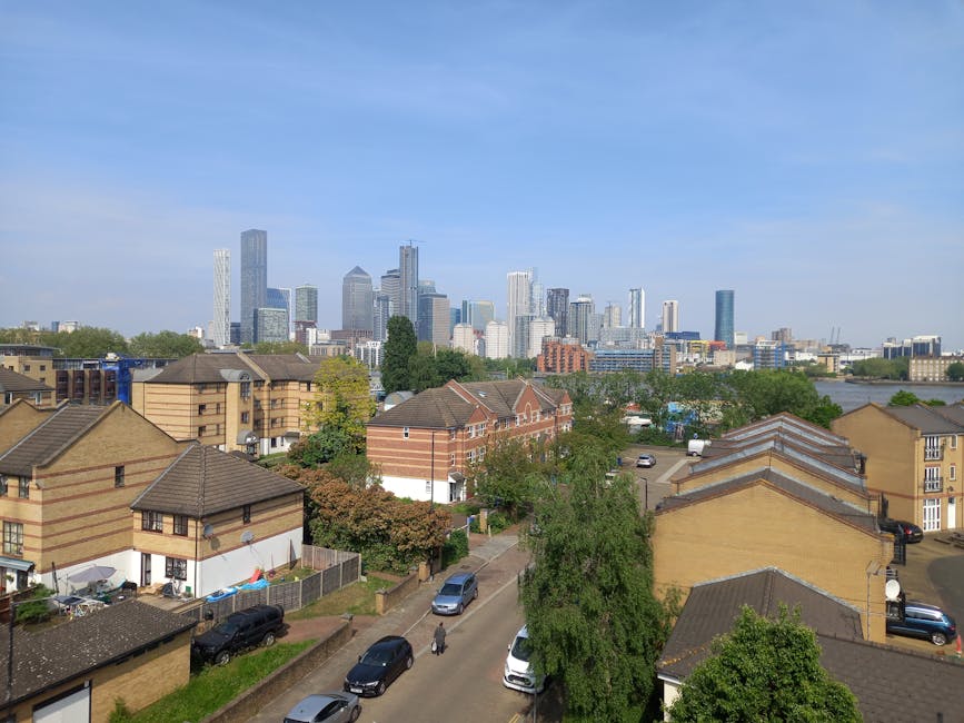 A panoramic view of Jubilee Park in Canary Wharf E14, showing residential houses with pitched roofs and brick exteriors in the foreground, surrounded by green trees and parked cars along the narrow streets. In the background, a cluster of modern skyscrapers with glass facades reflect sunlight under a clear blue sky. The scene highlights an urban residential area adjacent to a vibrant business district, with visible clean and well-maintained surfaces both outdoors and on building exteriors, suitable for deep cleaning and surface maintenance as part of domestic cleaning services from Cleaners E14.