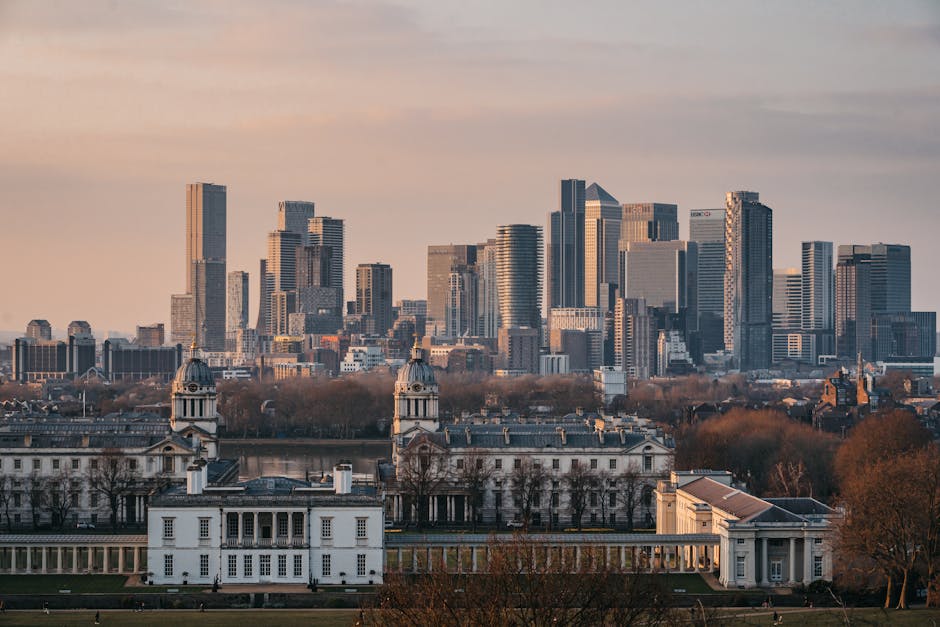 A panoramic view of Canary Wharf's modern high-rise office buildings with glass and steel facades reflecting the daylight, set against a soft pastel sky during sunset. In the foreground, the historic Queen's House, a white classical building with a symmetrical façade, domed towers, and columns, is visible surrounded by leafless trees and manicured lawns. The scene showcases a contrast between contemporary corporate architecture and classical heritage, with clear, well-maintained surfaces indicating a clean and tidy environment—a typical setting where professional cleaning services like those from Cleaners E14 are essential for maintaining hygiene and presentation in residential and commercial spaces.