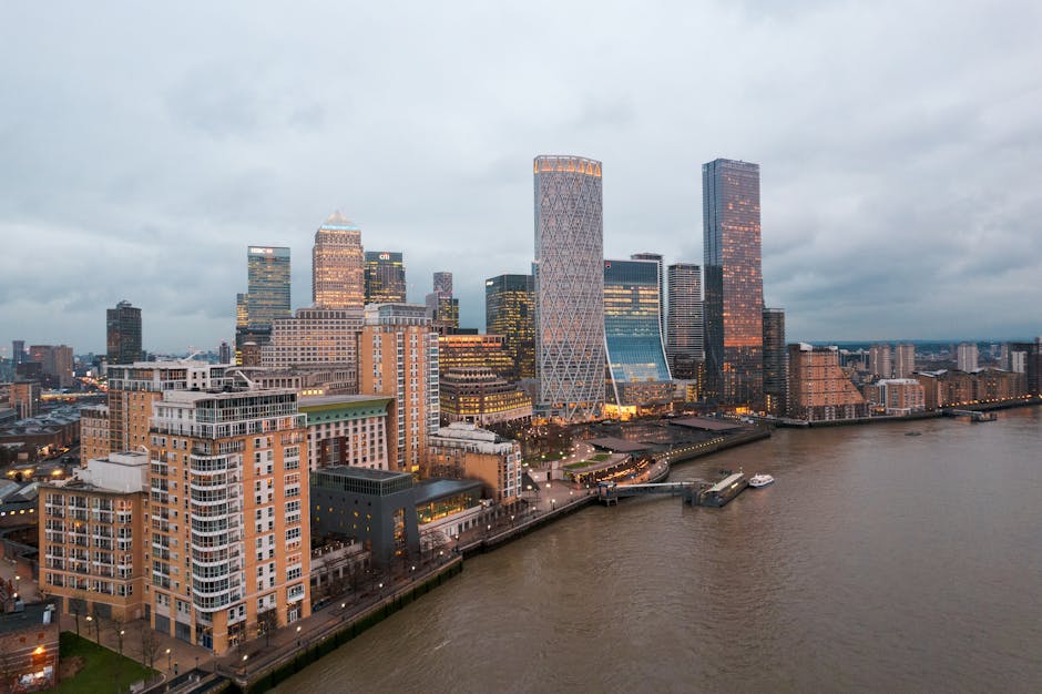 A panoramic view of Jubilee Park in Canary Wharf E14, showing residential houses with pitched roofs and brick exteriors in the foreground, surrounded by green trees and parked cars along the narrow streets. In the background, a cluster of modern skyscrapers with glass facades reflect sunlight under a clear blue sky. The scene highlights an urban residential area adjacent to a vibrant business district, with visible clean and well-maintained surfaces both outdoors and on building exteriors, suitable for deep cleaning and surface maintenance as part of domestic cleaning services from Cleaners E14.
