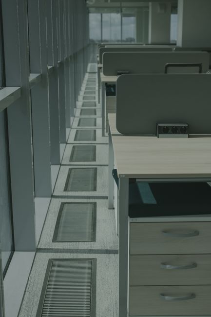 The image shows a modern office corridor with large windows allowing natural light to illuminate the space. Along one side, there are work desks with light-colored wooden surfaces, each equipped with a black power outlet and ergonomic grey partitions separating individual workspaces. The floor features a light grey, textured surface with built-in air vents or floor grilles positioned periodically along the corridor, indicating proper ventilation and hygiene maintenance. The corridor appears clean and well-maintained, with surfaces free of dust or debris. The setting suggests a professional environment, and the overall scene reflects a focus on surface cleaning and hygienic upkeep, consistent with office cleaning services provided by Cleaners E14 for workplaces on the Isle of Dogs.