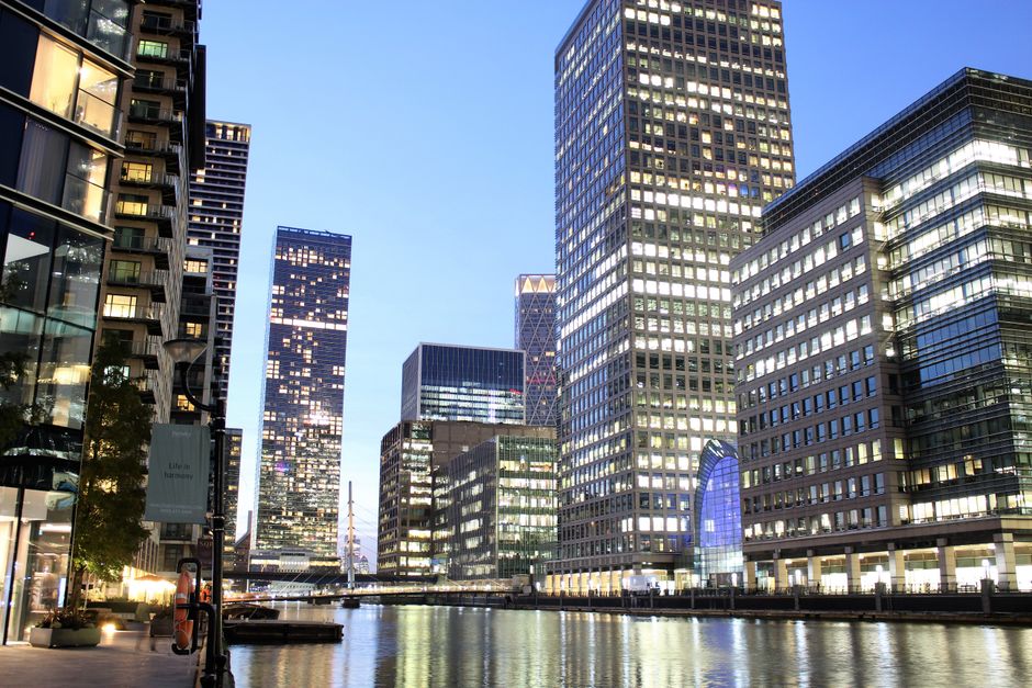 Image of a modern commercial office building at dusk with large glass windows reflecting the city lights, situated along a waterway. The building's exterior features a combination of clear glass and steel, with the interior lights creating a bright, clean appearance. Surrounding skyscrapers also have illuminated windows, showcasing an urban environment with a mix of steel, glass, and concrete surfaces. The scene includes a paved promenade with planters, trees, and street lamps, highlighting the urban setting typical of Isle of Dogs. As part of our cleaning services at Cleaners E14, this image illustrates the importance of maintaining pristine, hygienic office environments through surface cleaning and deep cleaning procedures, ensuring a professional and welcoming workspace.