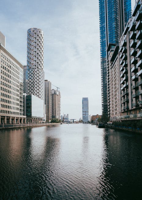 A high-angle view of a city canal featuring calm water reflecting the surrounding modern skyscrapers and office buildings. The buildings are made of glass, steel, and concrete, with some displaying patterned facades and large windows. The sky above is partly cloudy with soft sunlight illuminating the urban landscape. In the foreground, the canal’s surface appears wet and slightly rippled, while the edges are lined with waterfront walkways. This scene emphasizes the clean, urban environment typical of South Quay, which is often maintained by professional cleaning companies such as Cleaners E14 for residential and commercial properties. The image highlights the importance of surface cleaning and proper maintenance in such metropolitan settings.