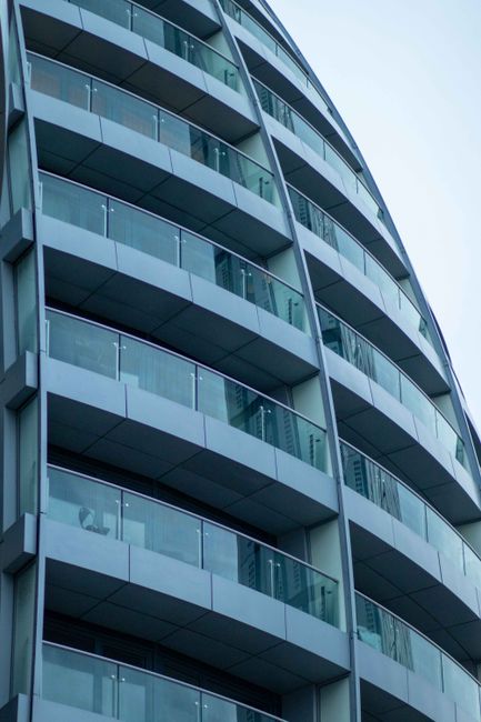 Close-up view of a modern residential apartment building with multiple curved balconies featuring glass railings, set against a clear blue sky. The building's facade consists of smooth, light-colored panels and large glass windows that reflect the surroundings. The balconies have clean, transparent glass panels, with some showing visible reflections and shadows. The overall appearance suggests a recently constructed, well-maintained structure. This image exemplifies external surface cleaning and maintenance for high-rise residential properties, similar to those in West India Quay, E14. For professional cleaning and surface maintenance of such buildings, [COMPANY_NAME] provides specialised services in the E14 area, including deep cleaning, window sanitisation, and ongoing upkeep to preserve architectural aesthetics and hygiene.