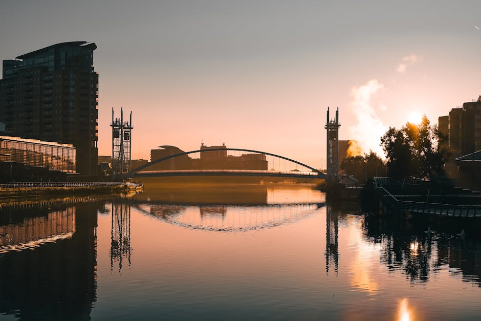 A calm river reflecting the surrounding modern buildings and a bridge in the early morning light, with a clear sky and warm sunlight illuminating the scene. The river surface appears clean and smooth, with visible reflections of the structures and trees along the banks. The scene exhibits a serene urban landscape, with some steam rising from a chimney on the right side, and the area is well-lit by natural sunlight, highlighting the neat and tidy environment. This image visually aligns with surface cleaning and maintenance themes, emphasizing cleanliness and hygiene in urban settings, as promoted by Cleaners E14 on their West India Quay apartment cleaning guide page for E14 residents.