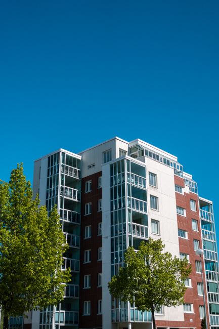 Close-up view of a modern residential apartment building with multiple curved balconies featuring glass railings, set against a clear blue sky. The building's facade consists of smooth, light-colored panels and large glass windows that reflect the surroundings. The balconies have clean, transparent glass panels, with some showing visible reflections and shadows. The overall appearance suggests a recently constructed, well-maintained structure. This image exemplifies external surface cleaning and maintenance for high-rise residential properties, similar to those in West India Quay, E14. For professional cleaning and surface maintenance of such buildings, [COMPANY_NAME] provides specialised services in the E14 area, including deep cleaning, window sanitisation, and ongoing upkeep to preserve architectural aesthetics and hygiene.